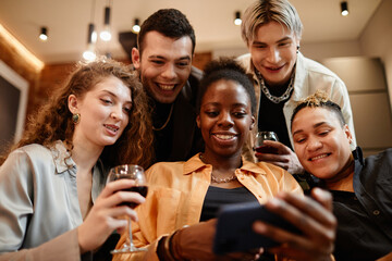Group of happy young intercultural friends gathered in living room looking through new photos in smartphone held by black girl