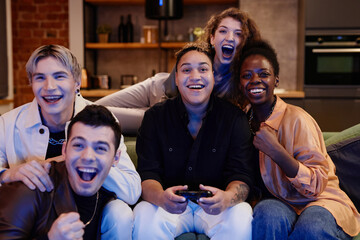 Group of young intercultural excited friends sitting in front of tv set and playing video game together while black woman using joystick