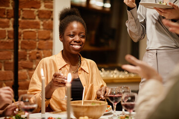 Young cheerful African American woman with glass of red wine enjoying home party while sitting by served table and chatting to friend