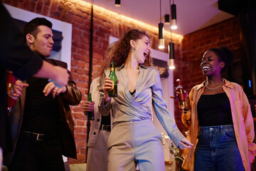 Happy young woman in grey suit with bottle of beer dancing among company of friends enjoying home party in living room