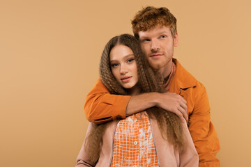 young redhead man hugging girlfriend with wavy hair isolated on beige.