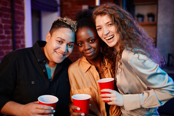 Three young cheerful intercultural women with alcoholic drinks standing in front of camera while enjoying home party in living room
