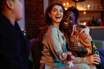 Fototapeta na wymiar Young laughing woman with bottle of beer embracing her girlfriend sitting next to her on couch and looking at another girl
