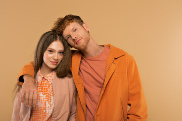 redhead young man in orange jacket hugging cheerful girlfriend isolated on beige.