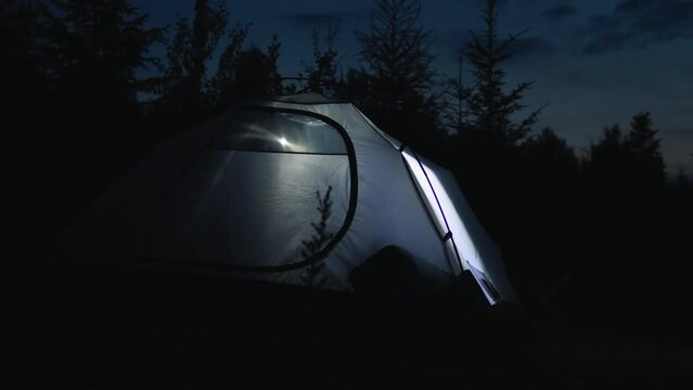 Tourist with flashlight in camp tent at night under sky. Tent at night with lights from flashlight. Gray tent lit from inside with flashlight against backdrop of amazing night landscape. Tourism
