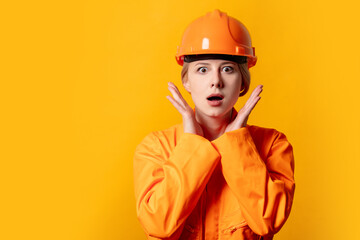 Surprised blond hair worker woman in orange helmet and suit on yellow background
