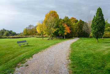 Fototapeta na wymiar Empty gravel footpath lined with benches and picnic tables leading through woods in a park under stormy sky in autumn