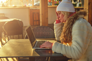 A woman in a sweater and a fur coat is sitting on the terrace of a cafe with a laptop. 