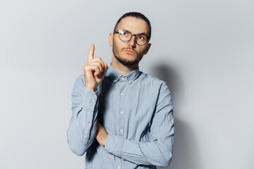 Studio portrait of young thoughtful man pointing index finger up, looking up, on white background. Wearing eyeglasses and blue shirt. New idea concept.