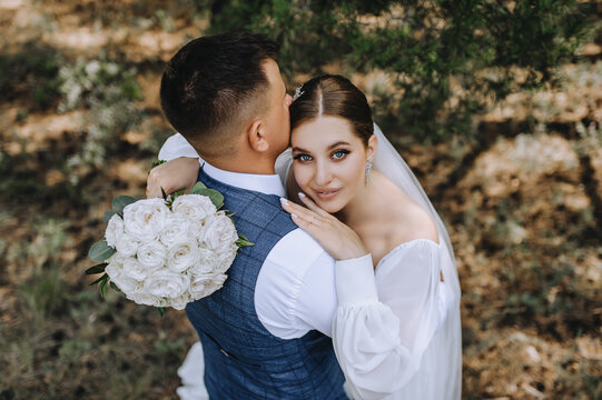 Beautiful newlyweds are hugging in the forest. Wedding portrait, photo of stylish groom and cute brunette bride with bouquet of roses.