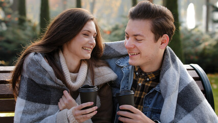 Joyful couple on romantic date in nature park covering with blanket keep warm talk carefree drink hot coffee herbal tea from disposable cups guy cuddle beloved girlfriend enjoy pleasant conversation