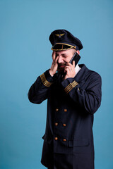 Middle aged pilot talking on smartphone with face palm expression, holding mobile phone, having conversation. Airlane captain in uniform standing, answering telephone call