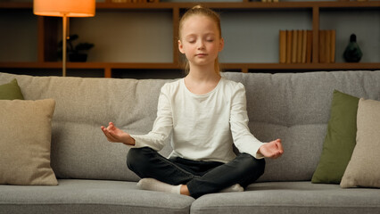 Little cute caucasian girl young healthy child blonde kid preschooler meditate sit on sofa at home...