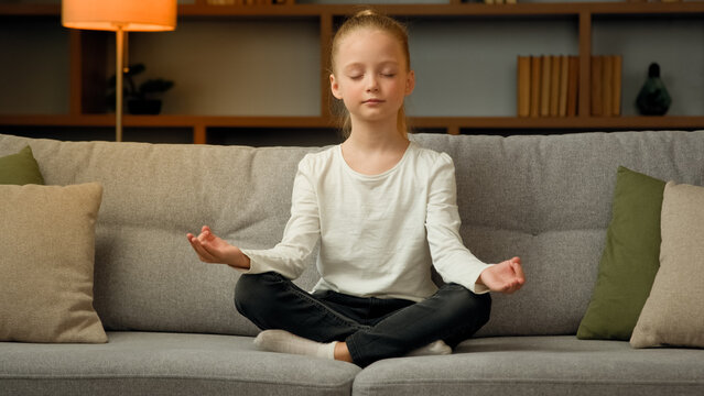 Little Cute Caucasian Girl Young Healthy Child Blonde Kid Preschooler Meditate Sit On Sofa At Home Calm Mindful Peaceful Daughter Do Yoga Breathing Exercise Relax Alone With Closed Eyes In Lotus Pose