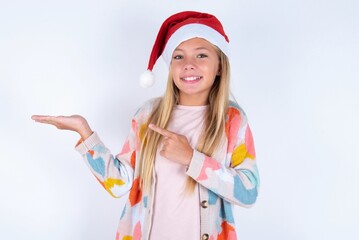 Fototapeta na wymiar little kid girl with Christmas hat wearing yarn jacket over white background pointing and holding hand showing adverts