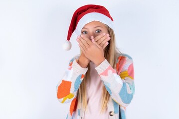 little kid girl with Christmas hat wearing yarn jacket over white background shocked covering mouth with hands for mistake. Secret concept.
