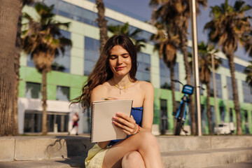 Beautiful fair-skinned young woman writing in notebook sitting in the stairs. On palms background holding pencil wearing blue top and yellow shorts. Studying outdoors concept 