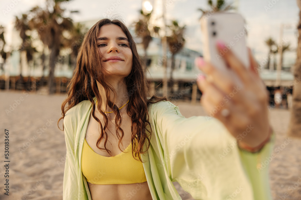 Wall mural european attractive cute woman smiling and looking to phone. doing selfie on the beach background. c