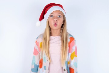 little kid girl with Christmas hat wearing yarn jacket over white background making fish face with lips, crazy and comical gesture. Funny expression.