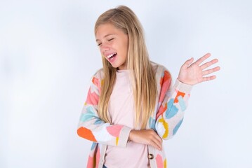 Overjoyed successful little kid girl wearing colorful yarn jacket over white background raises palm and closes eyes in joy being entertained by friends