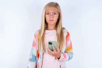 Upset dissatisfied little kid girl wearing colorful yarn jacket over white background  uses mobile software application and surfs information in internet, holds modern mobile hand