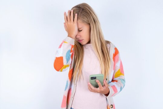 Upset depressed little kid girl wearing colorful yarn jacket over white background  makes face palm as forgot about something important holds mobile phone expresses sorrow and regret blames