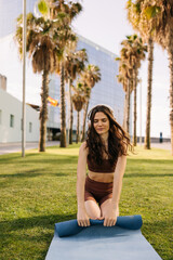 Picture of cheerful european woman twists yoga blue mat after workout. Sitting on green grass in headphones at park. Sport time outdoors, technology concept 