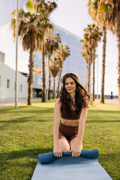 Picture of cheerful european woman twists yoga blue mat after workout. Sitting on green grass in headphones at park. Sport time outdoors, technology concept 
