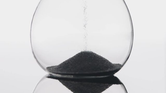 Grains of black sand and quartz are falling in a glass hourglass.