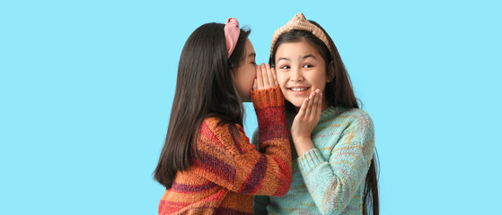 Cute little girl telling secrets to her sister on light blue background - obrazy, fototapety, plakaty