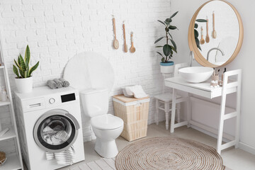 Interior of bathroom with modern washing machine, toilet bowl and sink near white brick wall