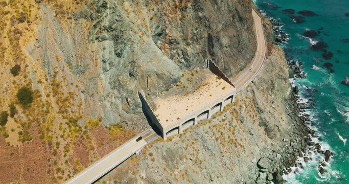 Cars moving by the dangerous road in the mountains of pacific shore. Turquoise ocean waves crush by the rocks. Top view.