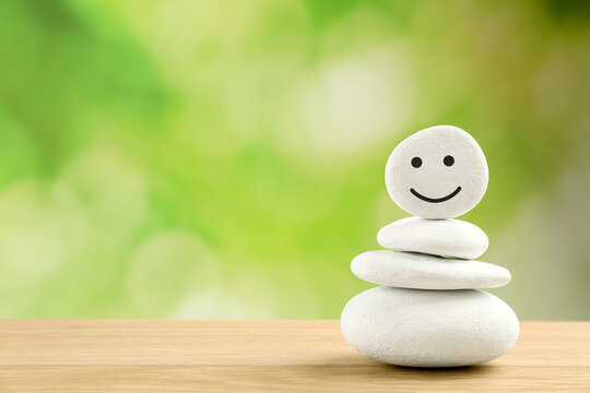 Stack of stones with drawn happy face on wooden table against blurred green background, space for text. Zen concept