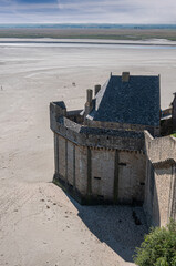 Mont St. Michel, Normandy, France - July 8, 2022: Gray-brown protruding corner section of rampart standing in ebb sandy beach with green marsh land in back and green tree line on horizon.