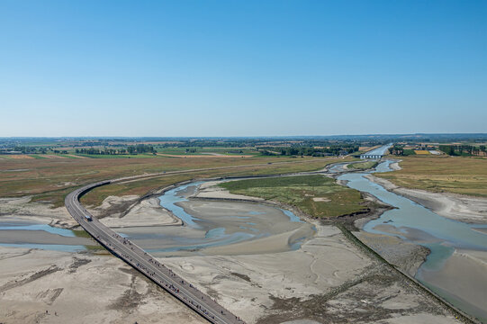 Mont St. Michel, Normandy, France - July 8, 2022: Seen from top of rock, sole approaching road with bridge over, and blueish Couesnon river cutting through sandy ebb beach under blue sky.