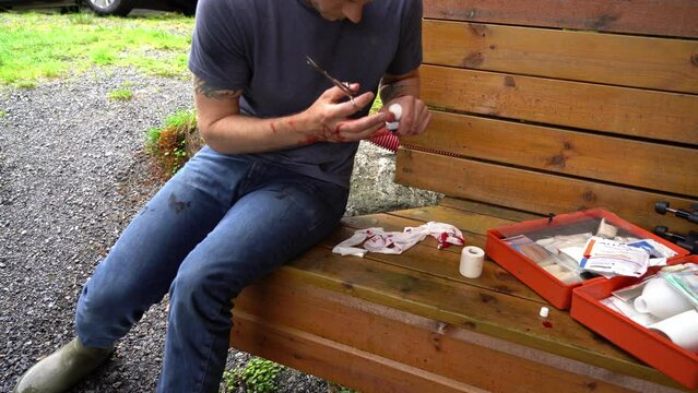 Man with injured hand is bleeding as he try to use first aid kit alone and bandaging his finger - Sitting outdoor on wooden bench with open first aid kit and blood spilled all over