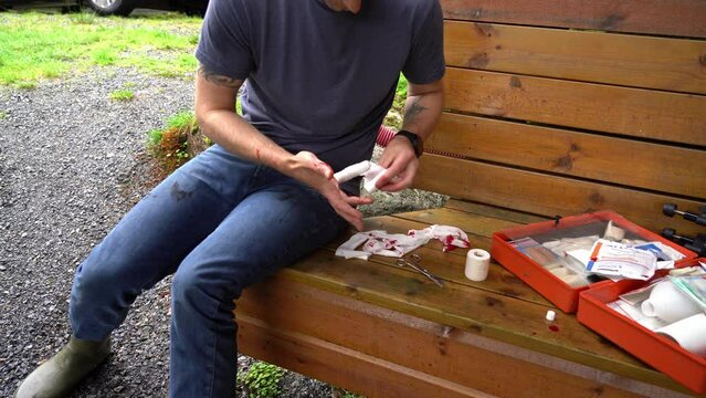 Male caucasian sitting outdoors on wooden bench and bandaging his bleeding finger - No face static clip with gravel background and open first aid kit to the right - Blood spilled on bandages and hand