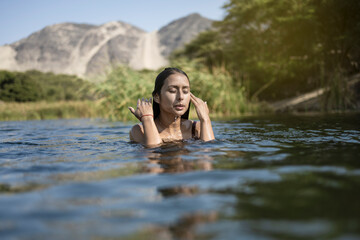 Beautiful young latin woman bathes in a lake enjoying a summer day. It stands at the bottom of the shoulders in the water. Her hands are raised above the water.