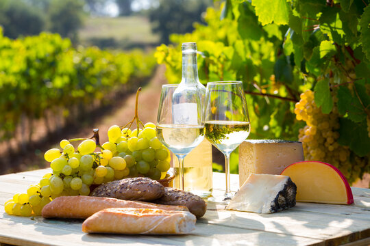Two wineglasses of white wine with baguette, various types of cheese on vineyard background