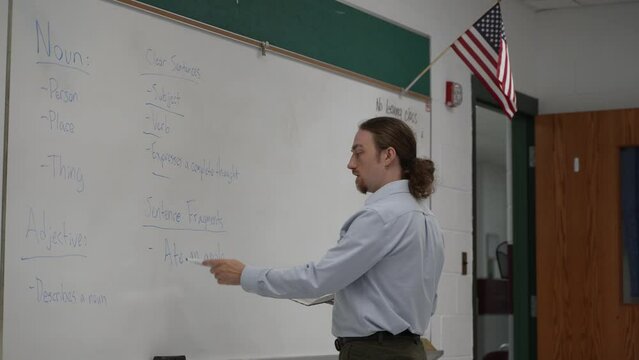 Male school teacher holding book teaching english in school classroom at a whiteboard, with US flag.