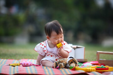 An Asian baby is sitting on a colorful mat eating a yellow block toy with other toys surrounding her in blur background.