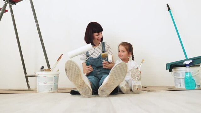 Cheerful caucasian woman mother and her cute little girl daughter sitting on floor in white light room background with paint rollers and brushes in their hands.