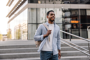 Modern young african man with backpack walks near city building. Dark-haired guy wears t-shirt, shirt and jeans. Lifestyle concept