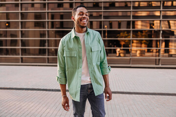 Modern young african man with wide smile laughs his eyes closed standing on street. Brunette guy with stubble wears casual clothes. Concept of enjoying moment