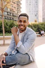 Fototapeta premium Smiling african young guy with snow-white smile looks at camera outdoors. Brunette man with stubble wears t-shirt and shirt. City life concept