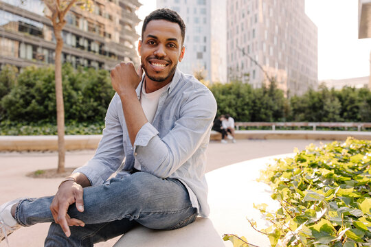 Stylish young african guy posing for camera sitting outdoors in city. Brunette man with stubble wears casual clothes. Relaxation concept