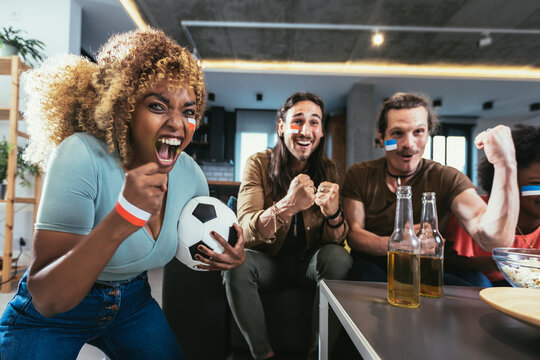Young group watching sports on television and cheering. Emotional football fans watching football game together at home. Sports Fans with Painted Faces Sitting on a Couch Watch Game on TV
