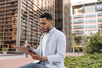 Focused young african guy is using tablet sitting on street. Man with dark hair wears shirt. Technology concept, lifestyle