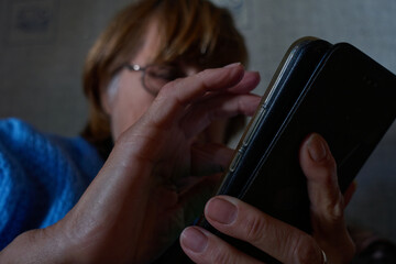 Grandmother dials a number on a smartphone. A red-haired old woman in glasses and a blue sweater sits on the sofa and looks into the phone. Selective focus. Dark photo.