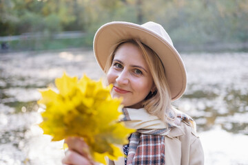 portrait of stylish woman in beige coat and hat holds autumn leaves in autumn park in fall